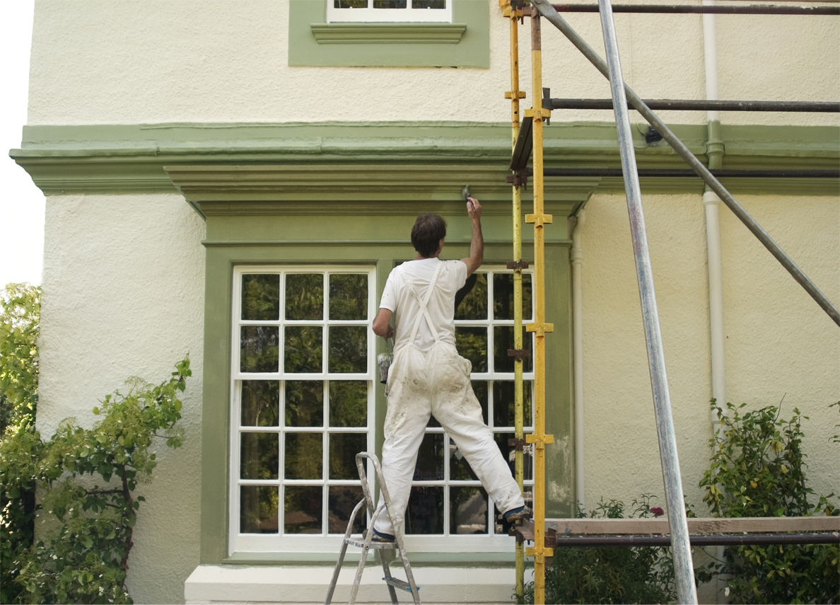 A man on scaffolding paints the frame of a house's window
