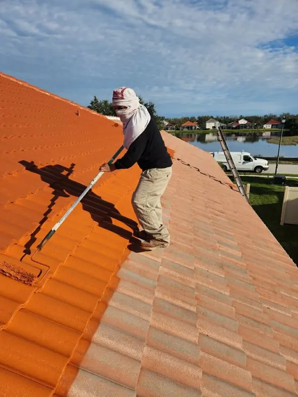 Painter rolling terracotta orange paint onto a tile roof at a lakefront home