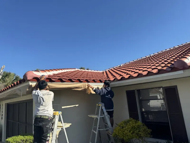Two Corrigan painters on ladders painting the fascia of a stucco home with a tile roof